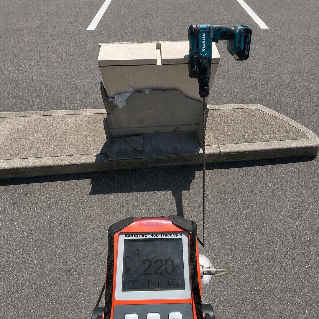 A tracer gas sensor being used in a parking lot with a drill to drill test holes.